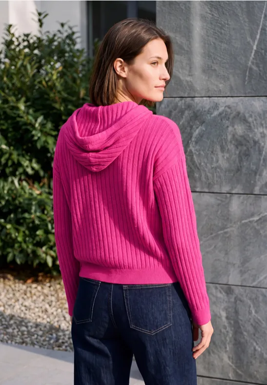 Vrouw van achteren met een roze capuchontrui en jeans, voor een stenen muur en struiken.