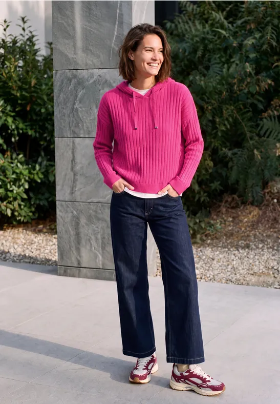 Vrouw lachtend in roze hoodie, donkerblauwe jeans en sneakers voor een stenen muur en groen loof.