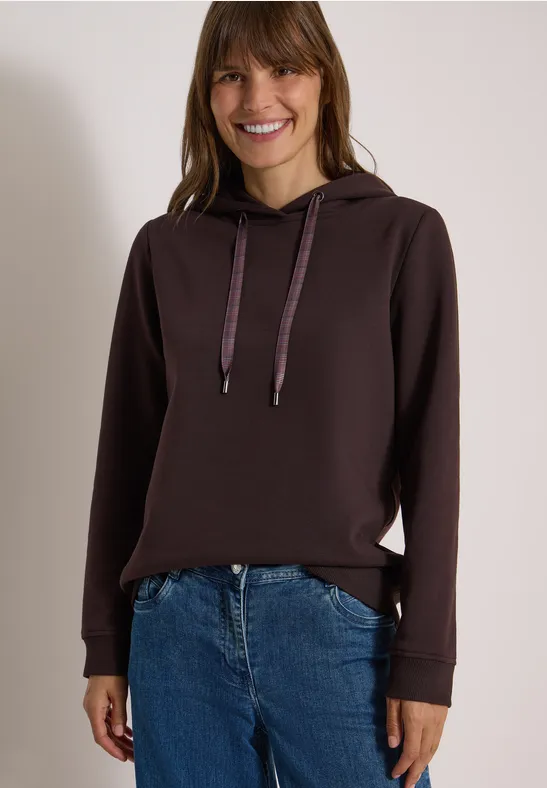 Eine lächelnde Frau trägt einen dunkelbraunen Kapuzenpullover und Jeans vor hellem Hintergrund.