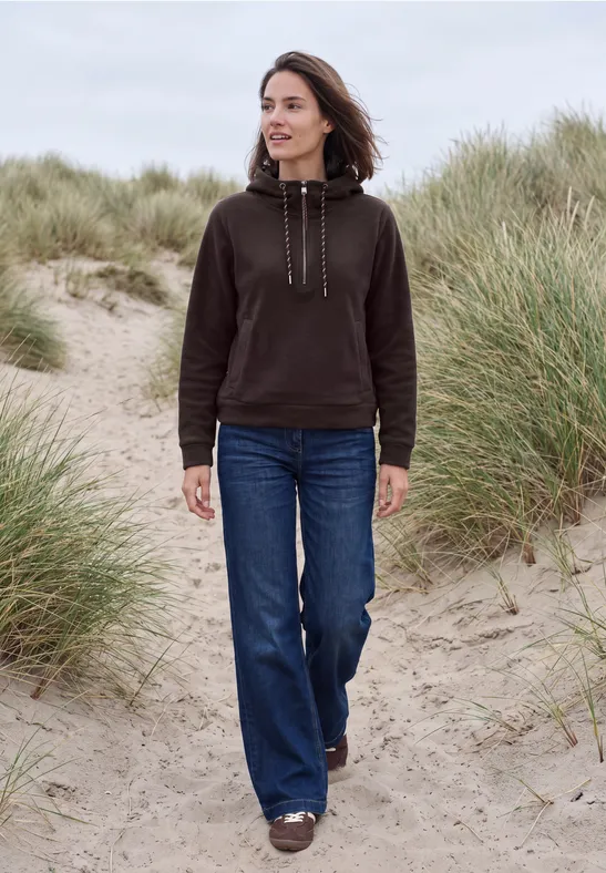 Frau im schwarzen Hoodie und blauen Jeans wandert über grasbewachsene Sanddünen am Strand.