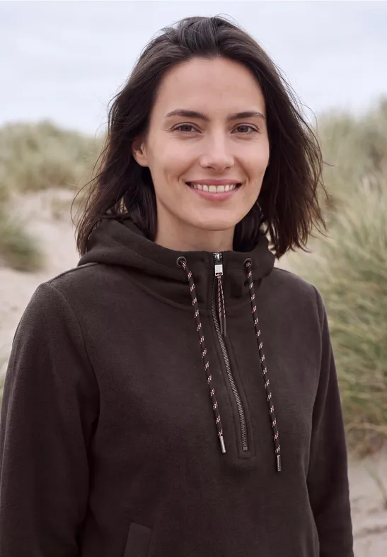 Lächelnde Frau in dunkelgrüner Kapuzenjacke stehend am Strand vor Dünen.