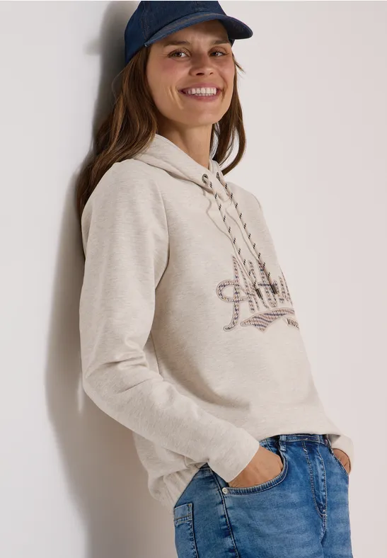 Eine lächelnde Frau in beiger Kapuzenjacke, Jeans und Basecap lehnt entspannt an einer Wand.