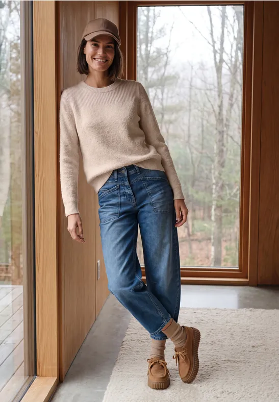 Een vrouw in een beige trui, spijkerbroek en mocassins lacht staand voor grote ramen in een modern houten interieur.