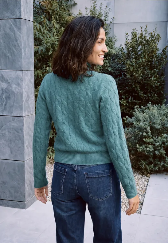 Femme de dos en pull vert et jean, souriante, se tient près de plantes devant un mur en pierre.