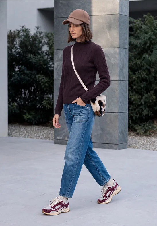 Femme en pull col roulé violet, jean et baskets marche avec sac à bandoulière devant un bâtiment moderne.