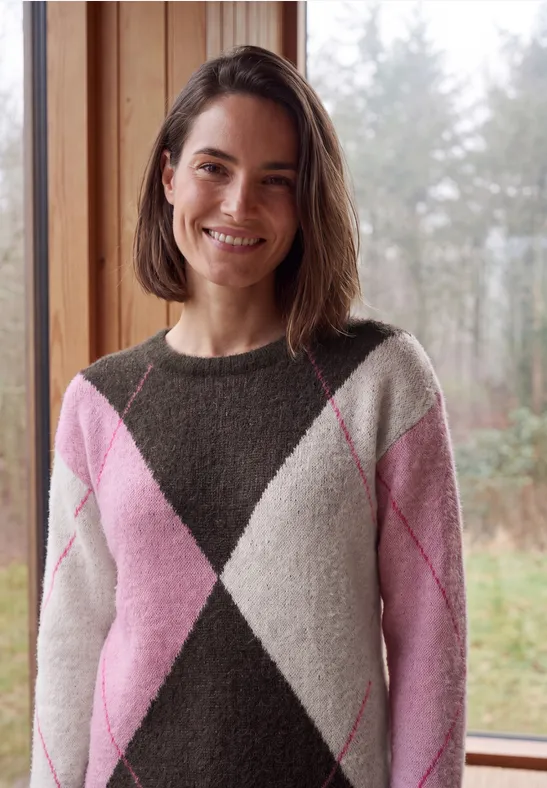 Frau mit schulterlangem Haar im rosa-weißen Rautenmuster-Pullover lächelt vor einem Fenster mit Waldblick.