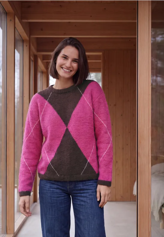 Lächelnde Frau mit rosa-braunem Rautenmuster-Pullover und Jeans steht in einem Holzrahmen und schaut in die Kamera.