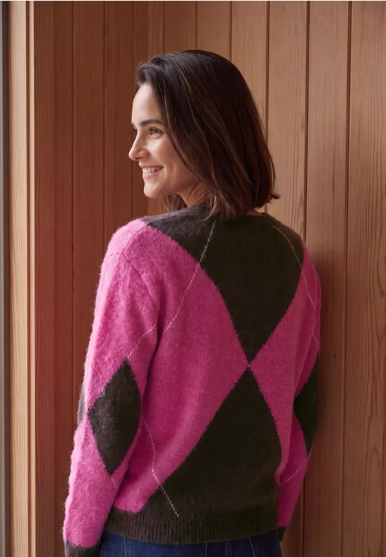 Frau lehnt am Fensterrahmen, trägt pink-grünen Rautenmuster-Pullover und lächelt zur Seite.