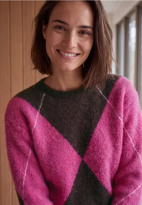 Eine lächelnde Frau mit schulterlangen braunen Haaren trägt einen pink-grün karierten Pullover vor Holzwand.