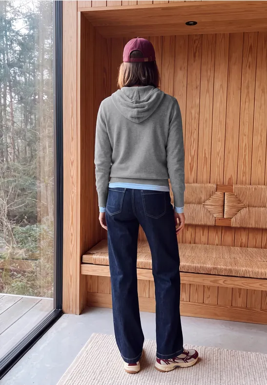 Une personne vue de dos porte un hoodie gris, un jean foncé et un bonnet dans une pièce en bois.
