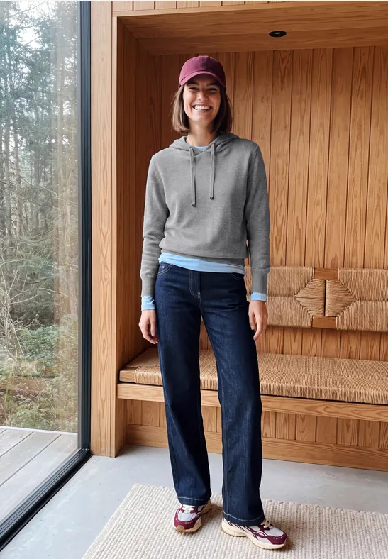 Personne en sweat gris, chemise bleue, jean foncé, baskets et casquette rouge dans un intérieur en bois