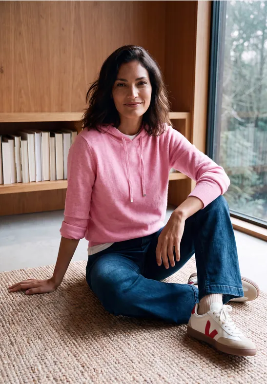 Frau in rosa Kapuzenpullover und Jeans sitzt auf Teppich vor Bücherregal.