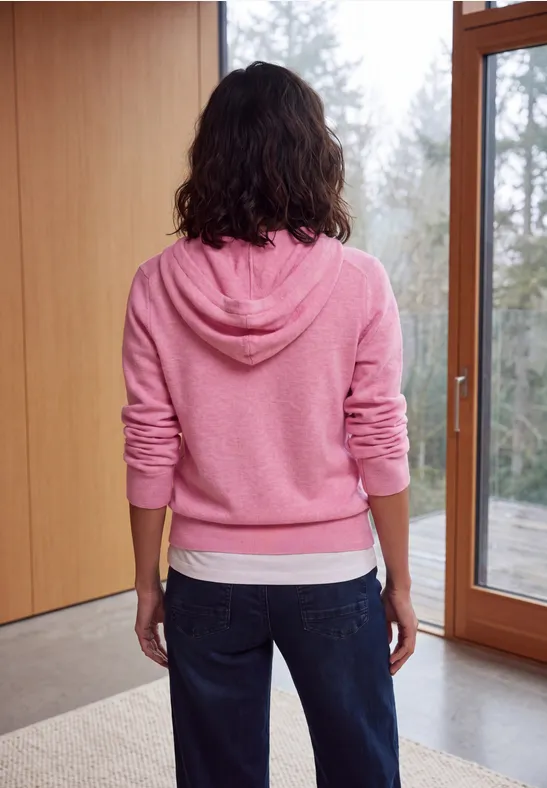 Frau von hinten in rosa Kapuzenpullover und Jeans steht vor großen Fenstern
