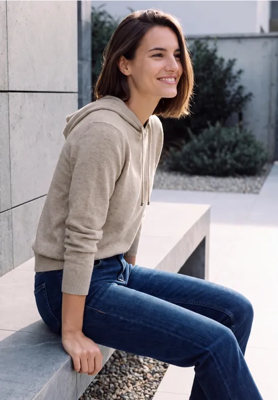 Eine junge Frau sitzt lächelnd auf einer Mauer, trägt Hoodie und Jeans, im Freien in modernem Umfeld.