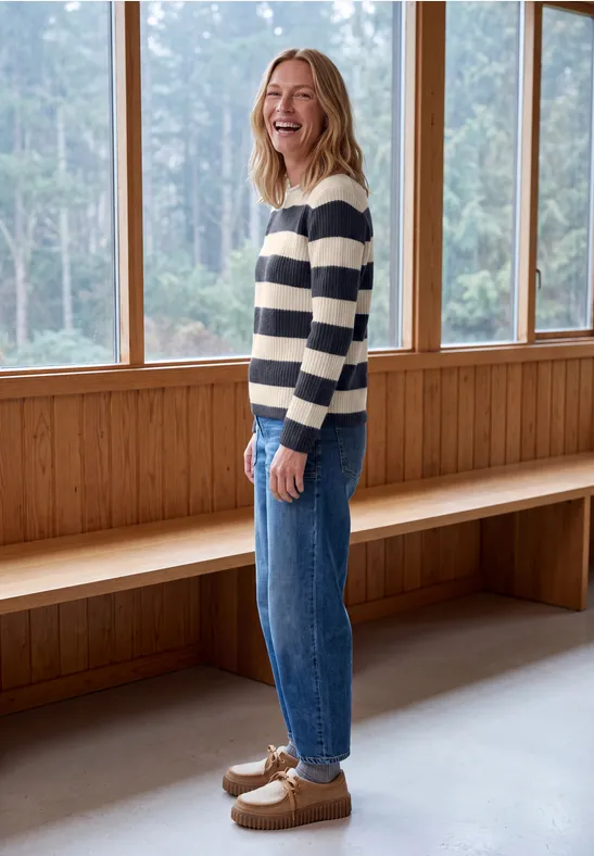 Une femme souriante en pull rayé et jean, debout devant des fenêtres et un mur en bois.
