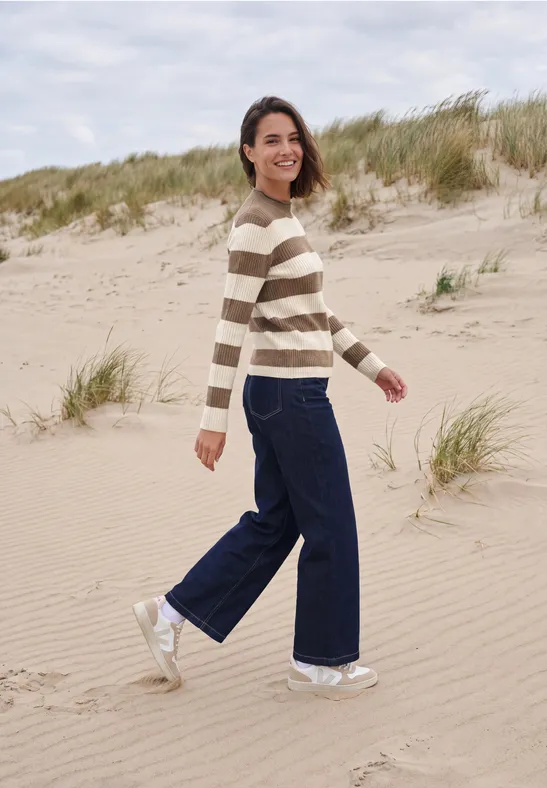 Femme souriante marchant sur la plage en pull rayé, jean large et baskets blanches.