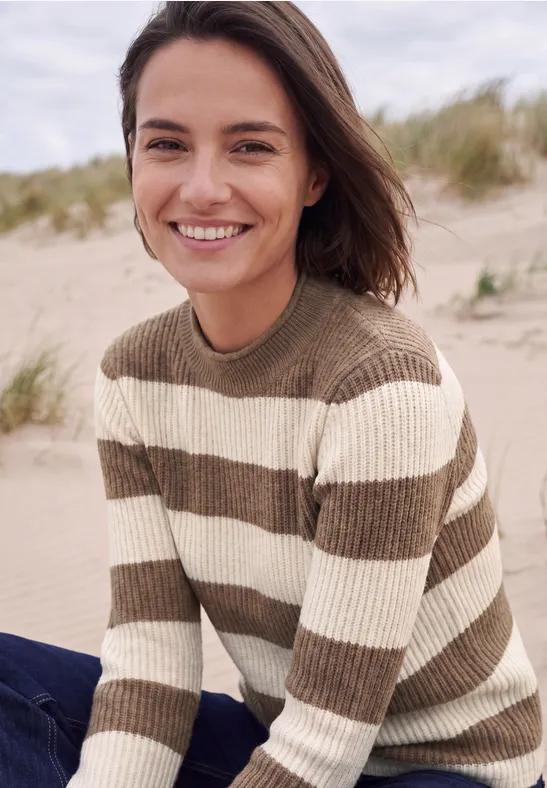 Une femme souriante en pull rayé sur une plage de sable.
