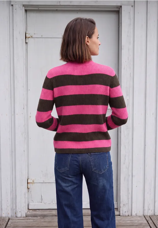 Une femme de dos porte un pull rayé rose et marron avec un jean bleu, devant une porte blanche.