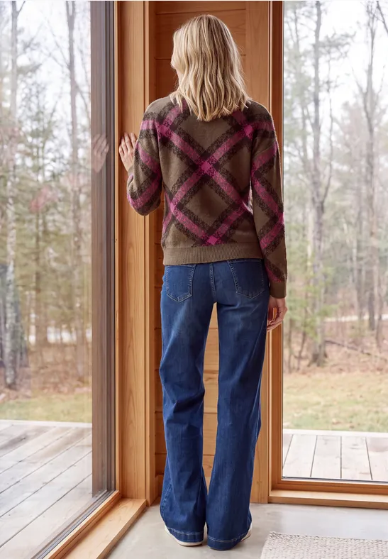 Frau in kariertem Pullover und weiten Jeans steht vor bodentiefem Fenster und blickt auf Wald.