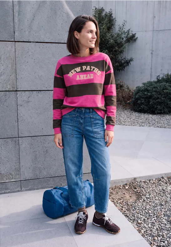 Een vrouw staat voor een betonnen wand, in een gestreepte trui, wijde jeans, loafers en tas naast haar.