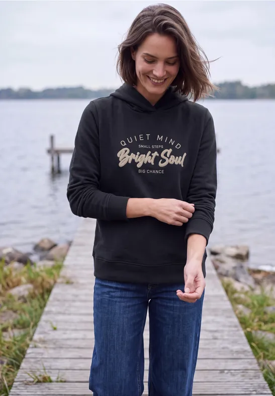 Eine lächelnde Frau in Jeans und schwarzem Kapuzenpullover steht auf einem Steg am Wasser.