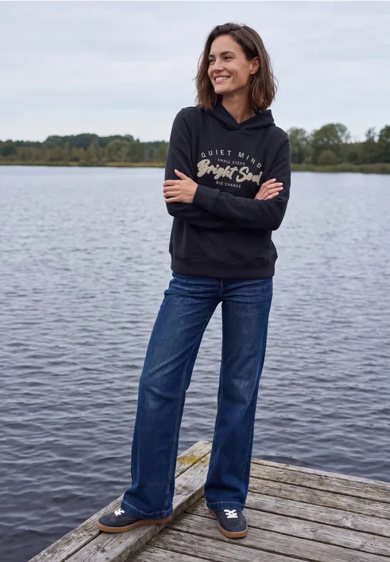 Eine Frau in schwarzer Kapuzenjacke und blauen Jeans steht lächelnd auf einem Holzsteg am Wasser.