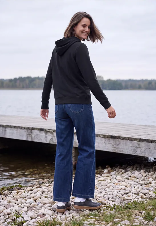 Eine Frau in schwarzer Kapuzenjacke und blauen Jeans steht am Ufer, blickt lächelnd zurück.