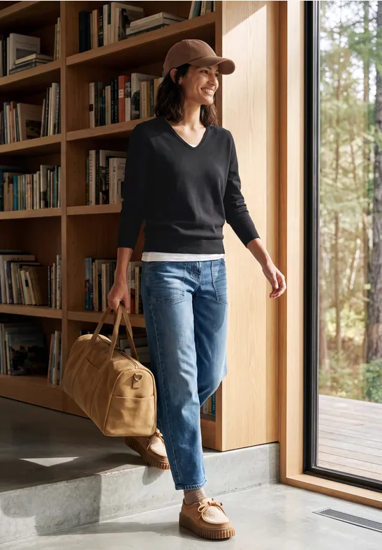 Frau mit Hut, Pulli und Jeans betritt mit Reisetasche ein modernes Holzhaus.