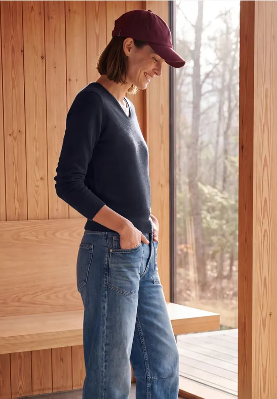 Frau in Jeans, dunkelblauem Pullover und Cap steht lächelnd seitlich vor einer Holzhütte im Wald.