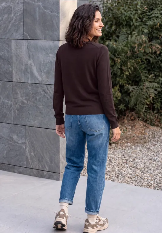 Femme vue de dos en pull marron et jean bleu marchant sur un chemin de gravier devant une maison moderne.