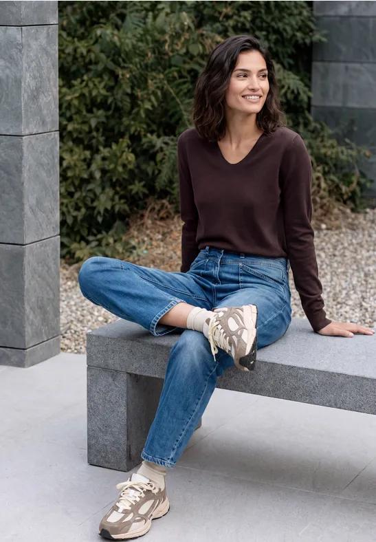 Une femme souriante est assise sur un banc en pierre à l'extérieur, porte un pull marron, un jean bleu et des baskets beiges.