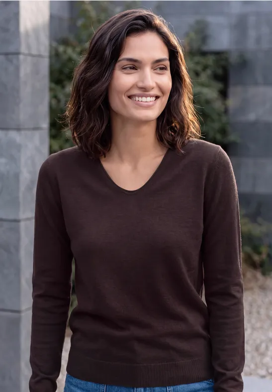 Une femme souriante aux cheveux foncés, portant un pull marron, posant devant un mur de pierre en plein air.