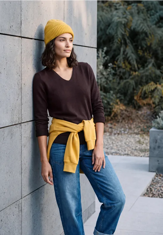 Femme appuyée contre un mur de béton, pull marron, jean, bonnet jaune et sweat noué à la taille.