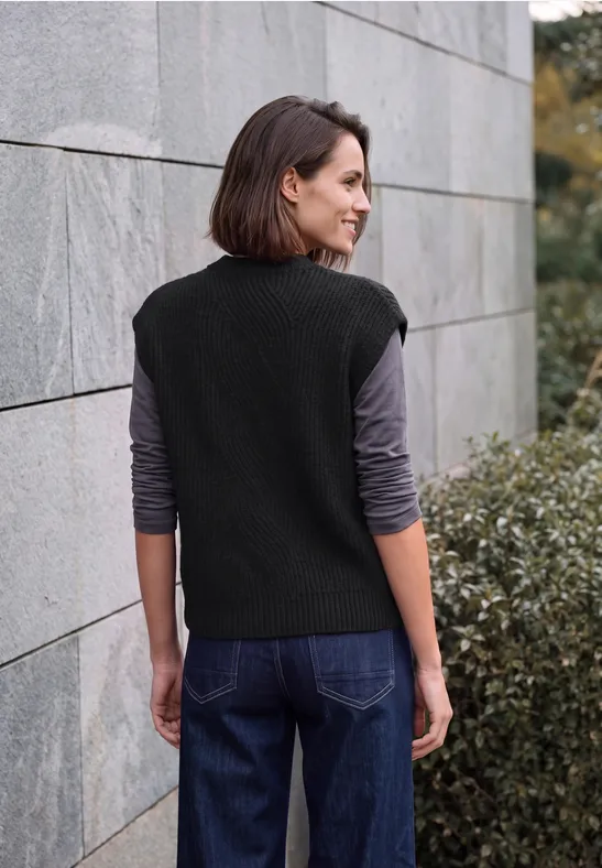 Frau von hinten in schwarzem Strickpullunder und langärmeligem Shirt neben einer Steinmauer und Sträuchern.