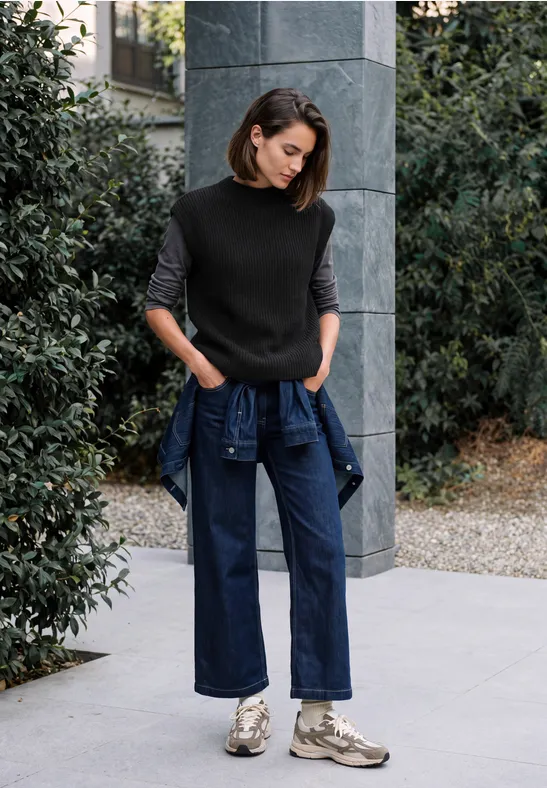 Eine Frau in schwarzem Pullover und weiten Jeans mit um die Hüfte gebundener Jacke steht draußen vor einer Säule.