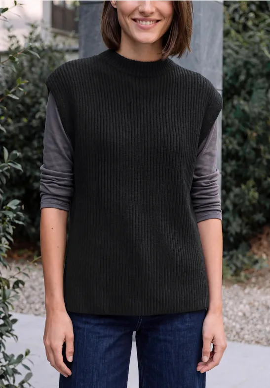 Frau mit schwarzer Strickweste über grauem Langarmshirt und Jeans, stehend vor einem Büschehintergrund.