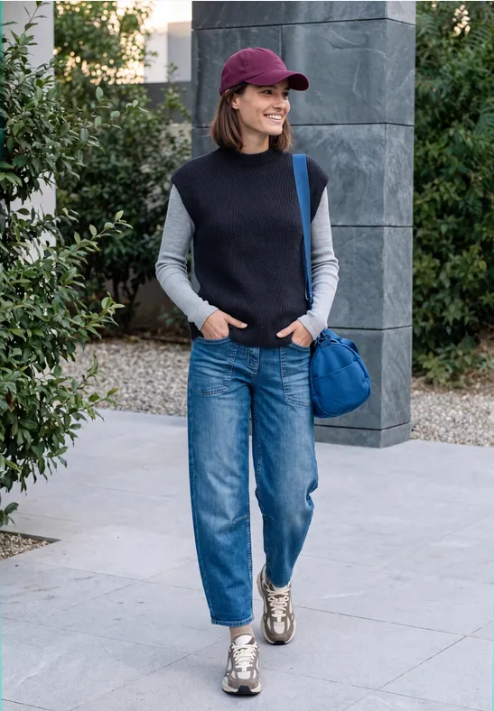 Frau in Jeans, Weste und Cap läuft mit blauer Tasche und Sneakers draußen spazieren.