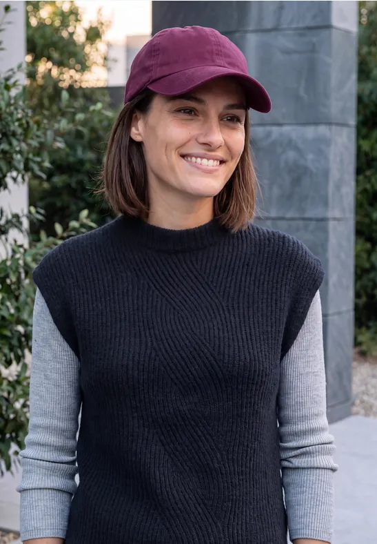 Eine lächelnde Frau mit lila Cap und schwarzem, ärmellosem Pullover über grauem Langarmshirt im Freien.
