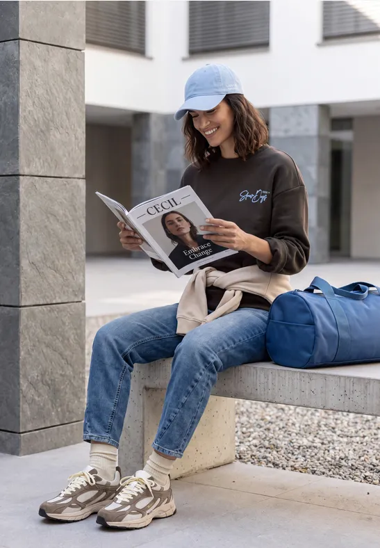 Een vrouw met pet leest rustig een tijdschrift bij een modern gebouw, naast haar blauwe reistas.