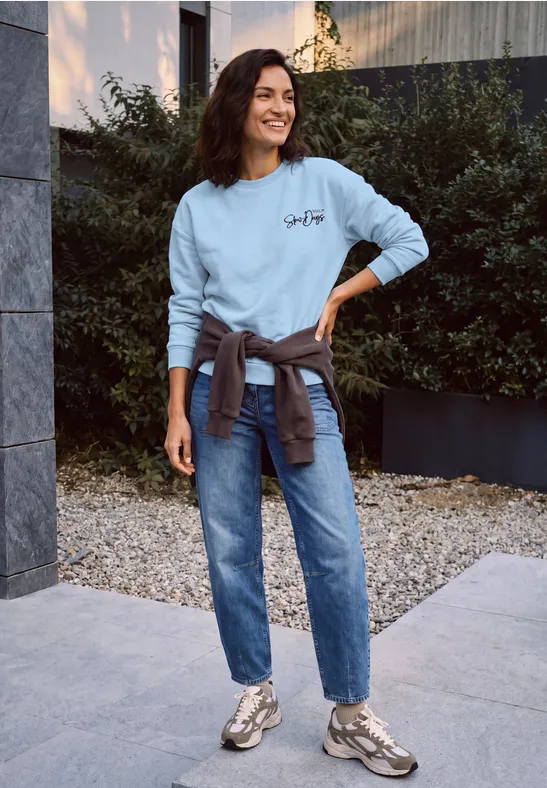Frau in hellblauem Sweatshirt, Jeans und Sneakern steht lässig auf einem Gehweg im Freien.