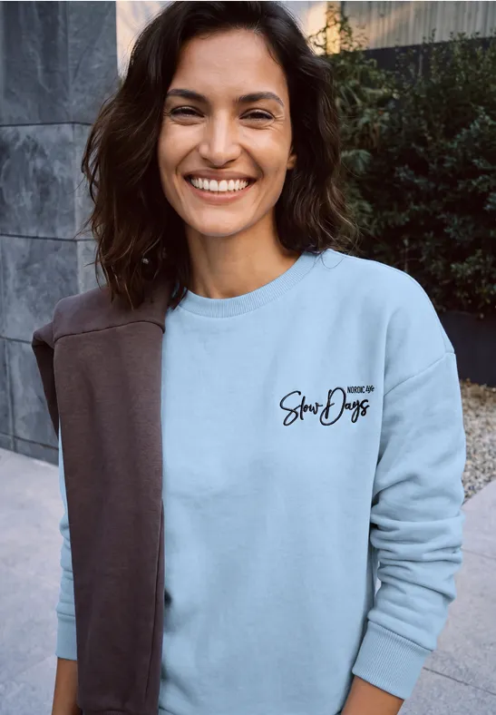 Frau lächelnd in hellem Pullover mit zweifarbigem Design und Schriftzug.