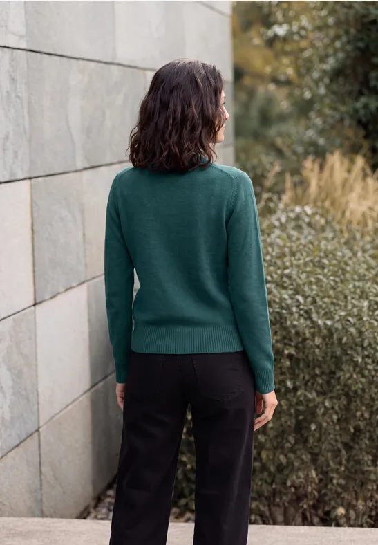 Femme en pull vert et pantalon noir, dos tourné, devant un mur de pierre avec des buissons.