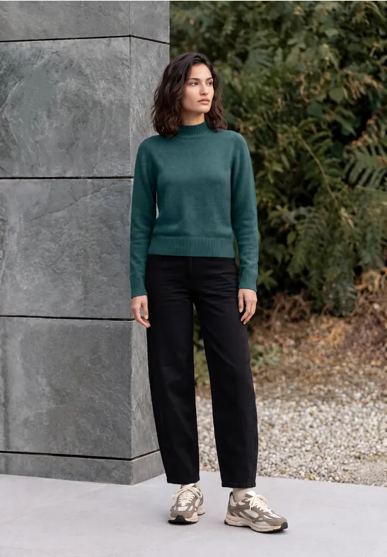 Femme décontractée en pull vert, pantalon noir et baskets, debout devant un mur de béton gris en plein air.