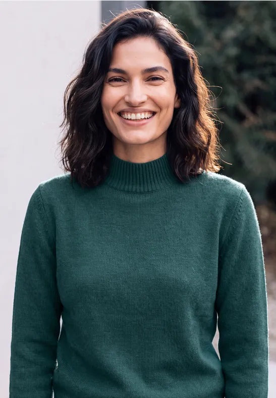 Une femme souriante aux cheveux bruns portant un pull vert devant un arrière-plan flou en plein air.