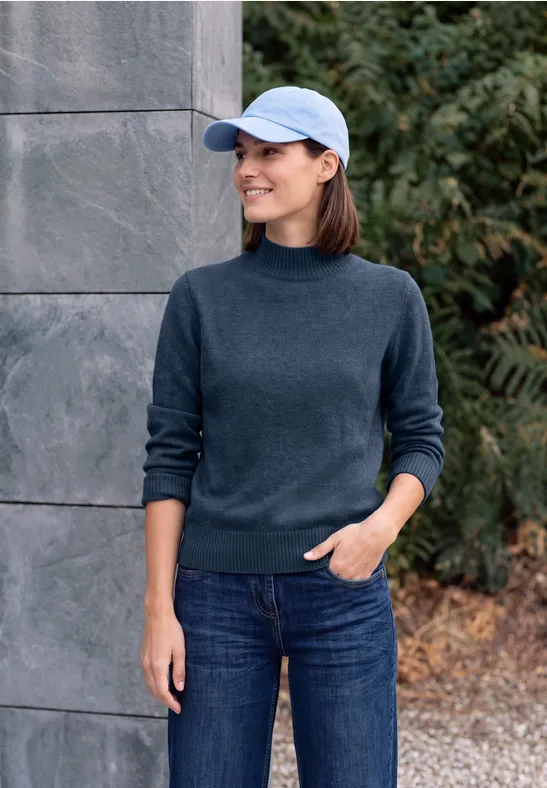 Femme en pull bleu foncé, jean et casquette, debout souriante devant un mur de pierre et des buissons.