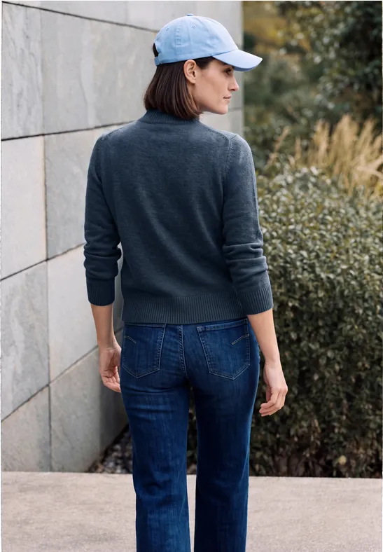 Femme en pull gris, jean et casquette, dos tourné, debout dehors près d'un mur et de buissons.