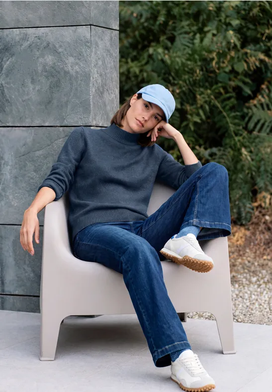 Femme détendue assise en extérieur sur un fauteuil beige, en jean, pull, baskets blanches et casquette bleue.