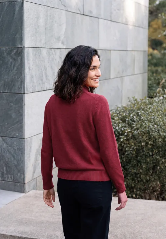 Femme en pull rouge près d’un mur, regard par-dessus l’épaule.