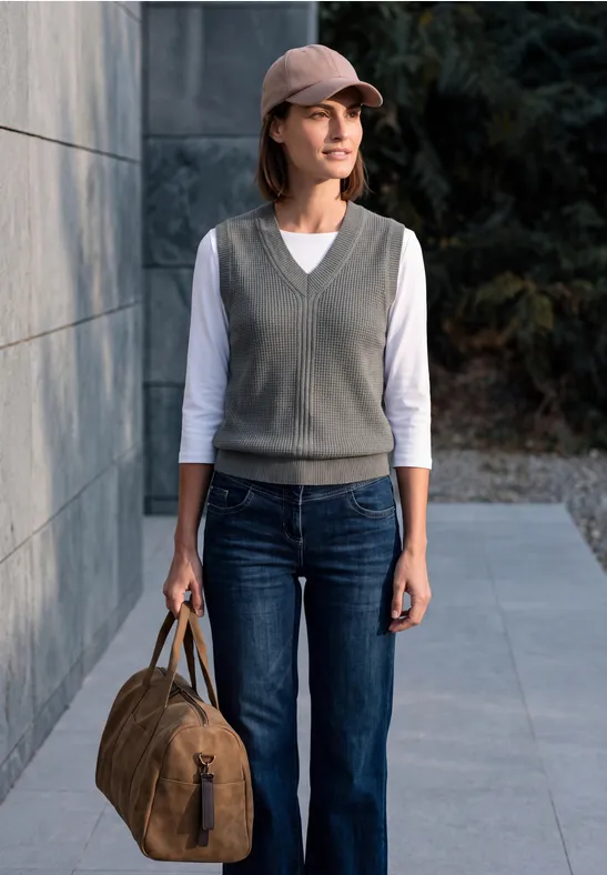 Frau in Jeans, grauer Weste und Cap steht mit brauner Reisetasche vor einer Hauswand.
