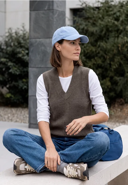 Frau mit Baseballkappe, Jeans und Weste sitzt im Freien auf einer Mauer, Tasche neben ihr.